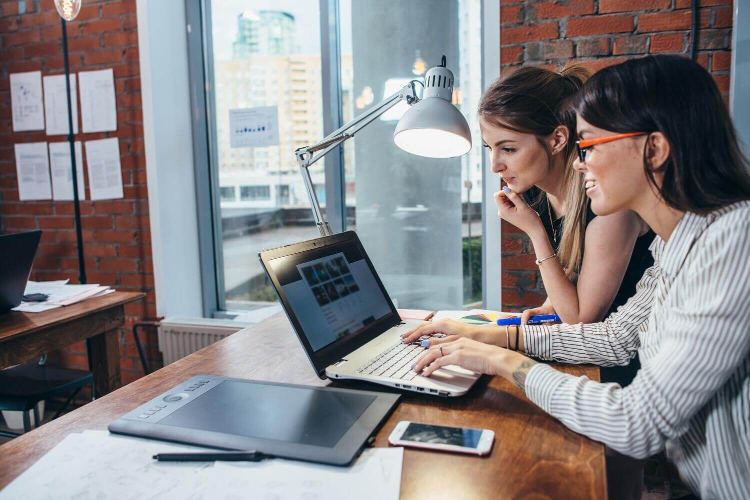 Two women collaborating on a laptop in a modern office setting, one typing and the other looking at the screen