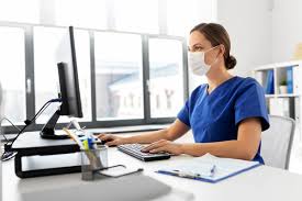 A healthcare professional wearing a face mask and blue scrubs sits at a desk, looking at a computer monitor and typing on a keyboard.