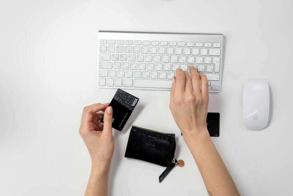 A person's hands are shown from above, holding a black digital camera in one hand and operating a white computer keyboard with the other hand, on a white surface.