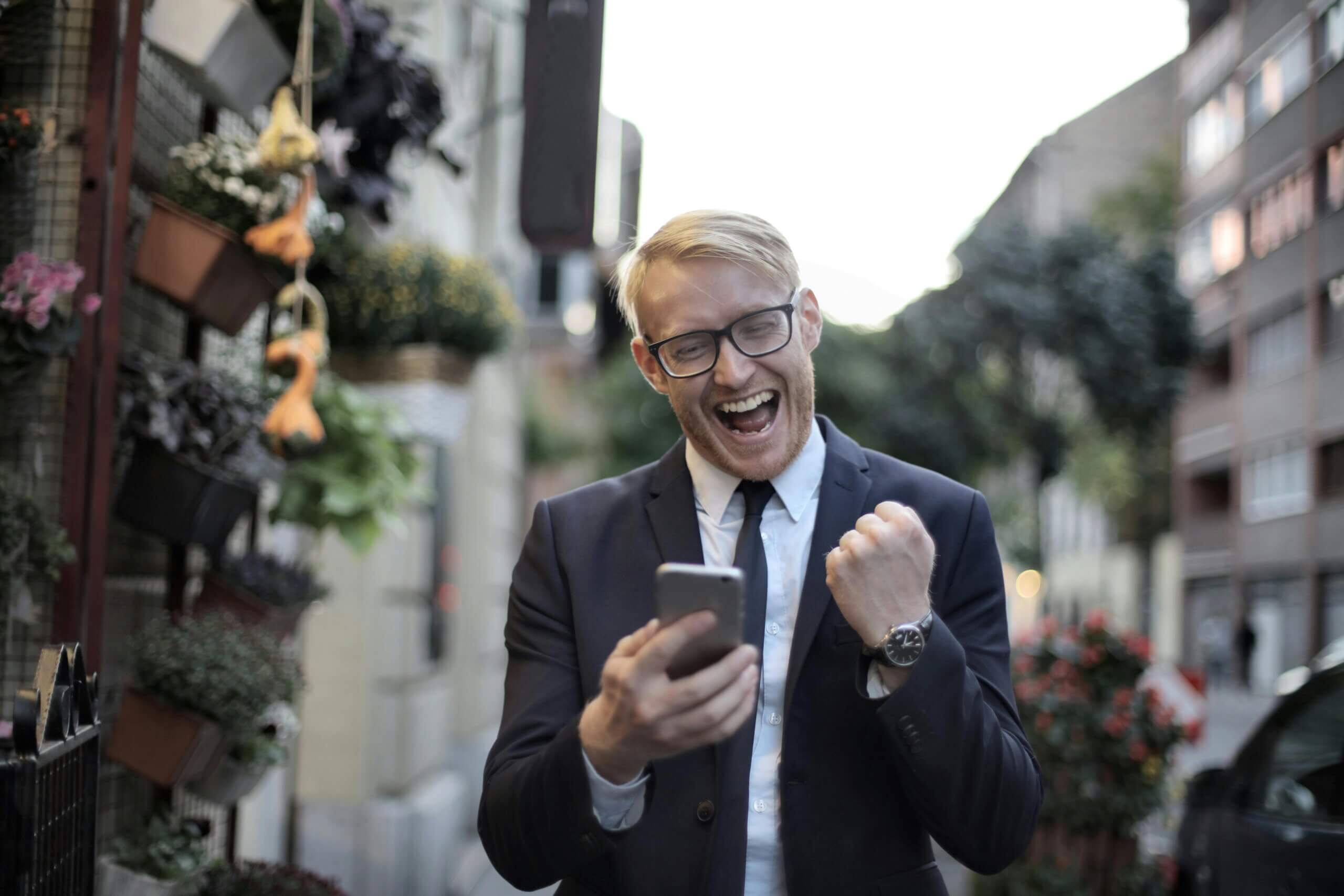 A happy businessman in a suit and glasses checks his smartphone on a city street, pumping his fist in celebration.