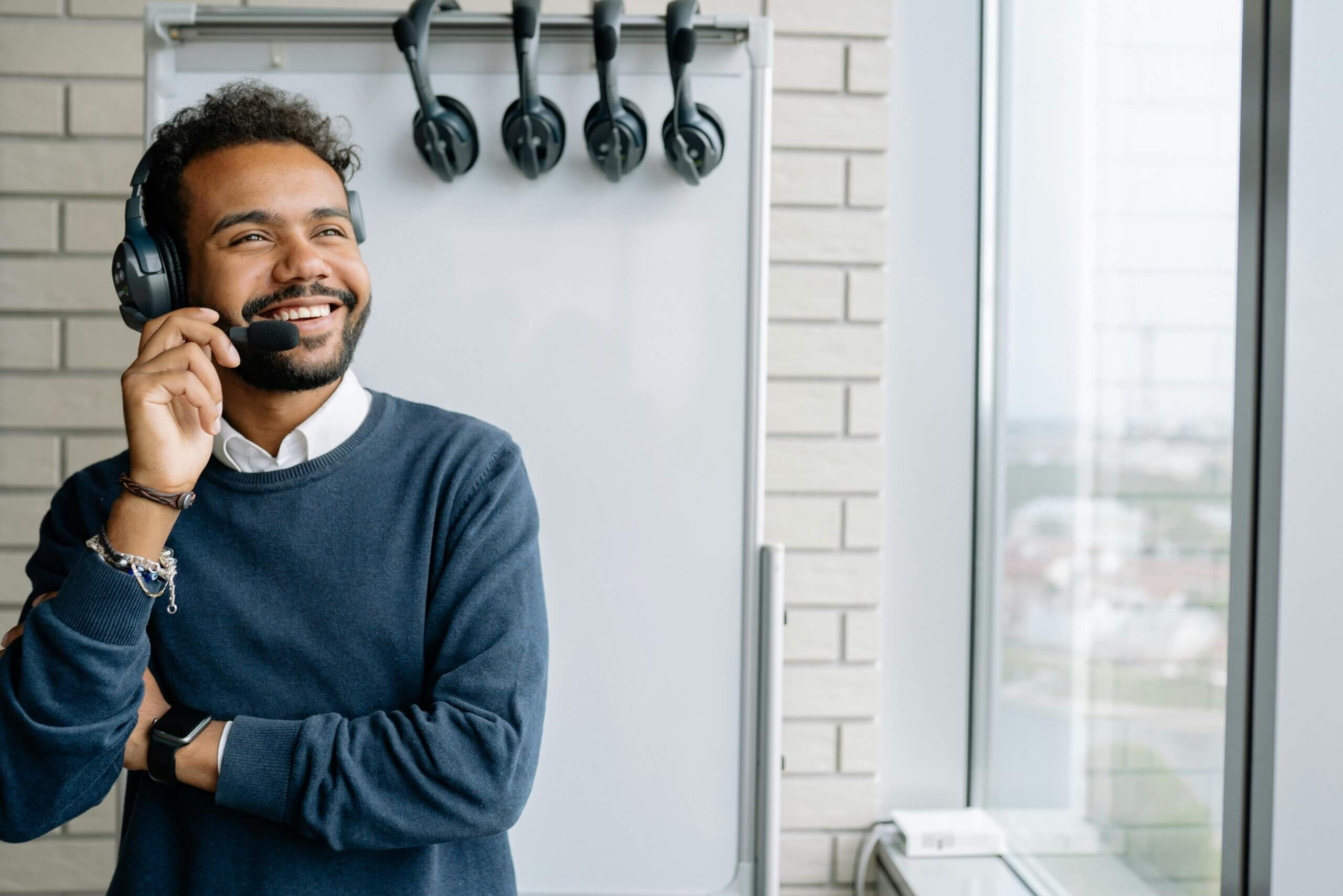A happy man with a beard wearing black headphones and a blue sweater, smiling while looking up and to the right, possibly in a call center or office setting.