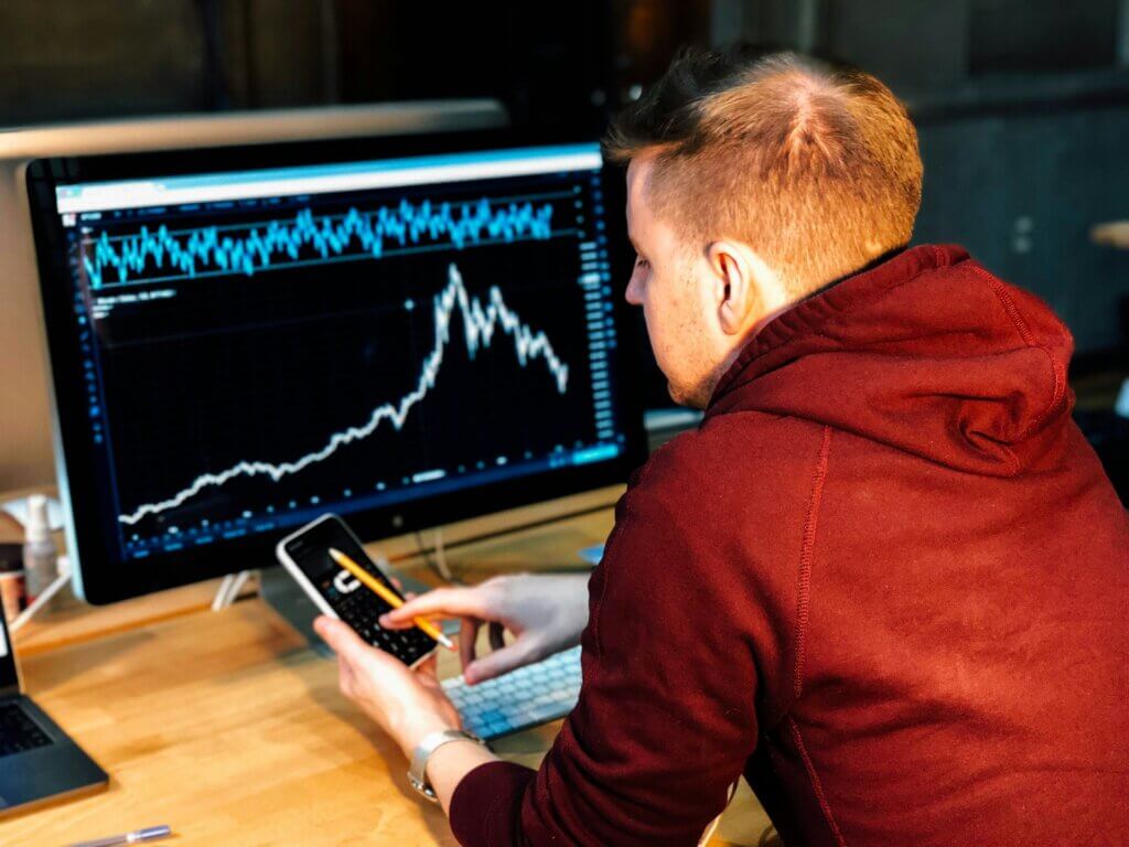 A person wearing a red hoodie sits in front of a computer monitor displaying financial charts, while simultaneously interacting with a smartphone showing a similar chart.